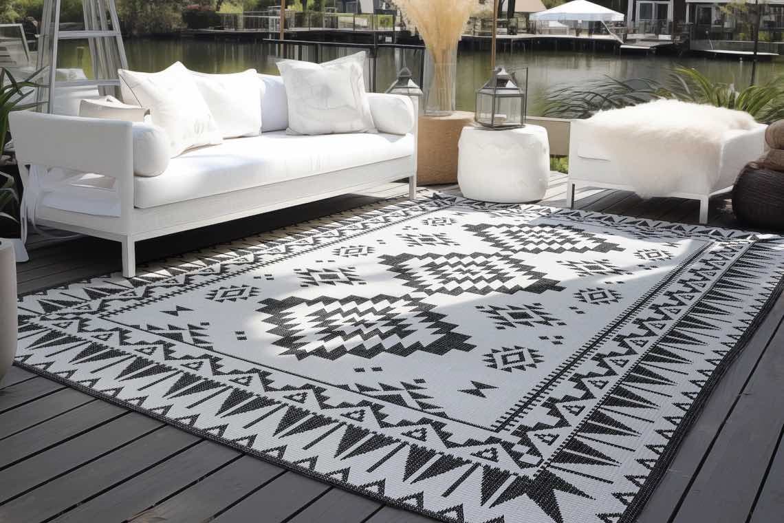 Terrasse mit Sofa, Kissen und dekorativem Teppich mit geometrischen Mustern, Blick auf Wasser und Boote im Hintergrund.