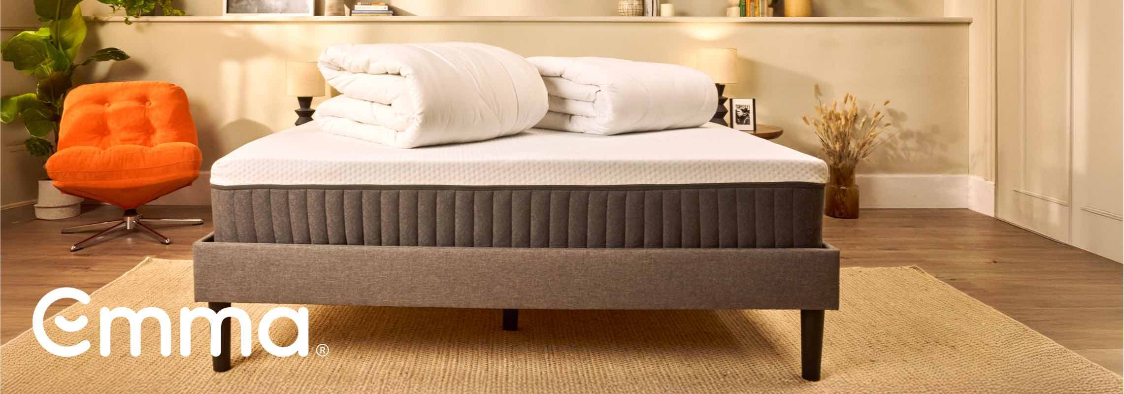 Emma Logo – graues Bett mit weißer Matratze und zwei gefalteten Bettdecken, orangefarbener Sessel, Zimmerpflanze im Hintergrund