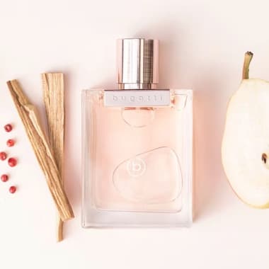 Bugatti Parfumflakon in Rosa, daneben Birnenhälfte, Holzstäbchen und rote Beeren auf hellem Hintergrund