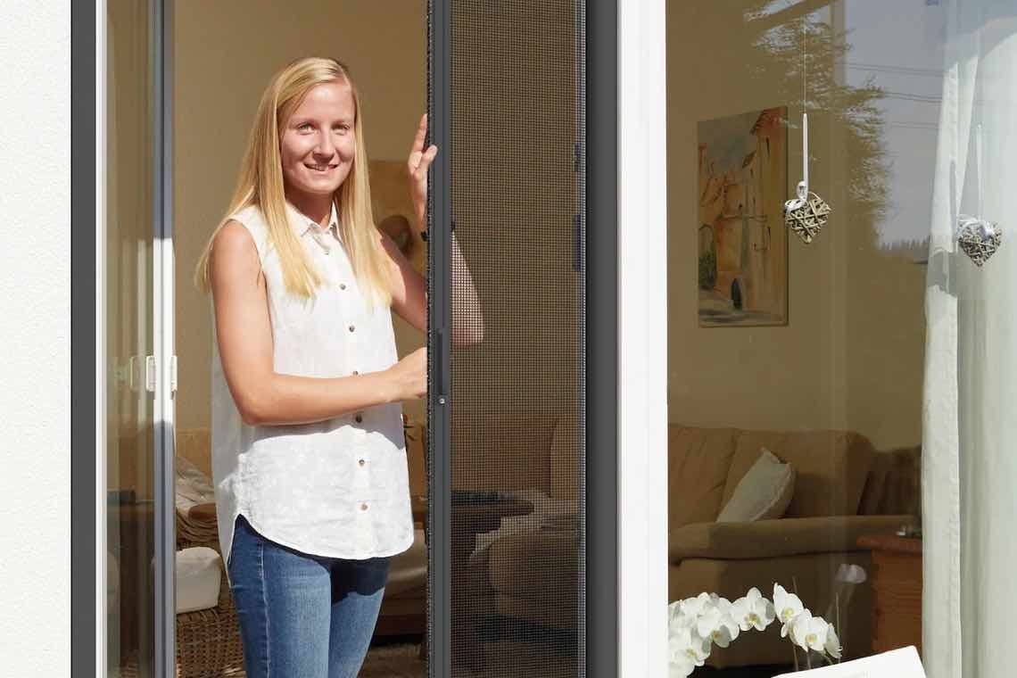 Frau öffnet eine Terrassentür mit Fliegengitter, dahinter Wohnzimmer mit Sofa und Deko-Herzen am Fenster