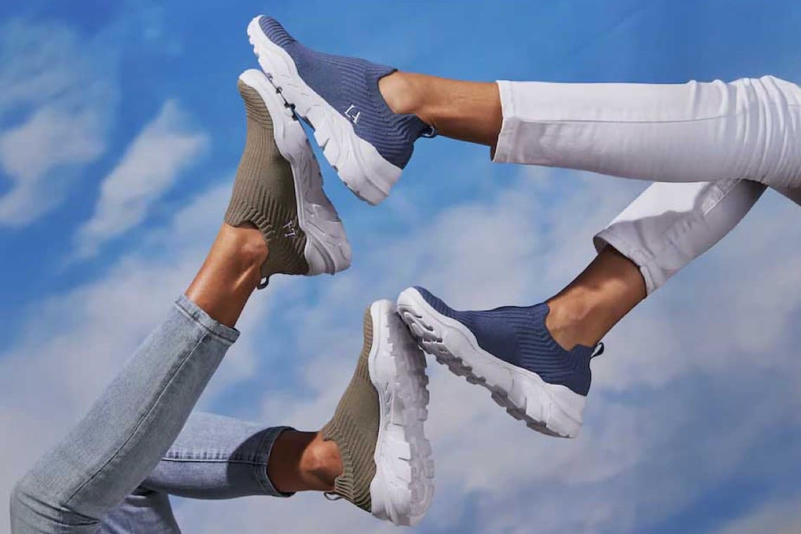 Vier Beine in Jeans strecken sich in die Luft, zeigen beige und blaue Slip-on-Sneaker vor blauem Himmel