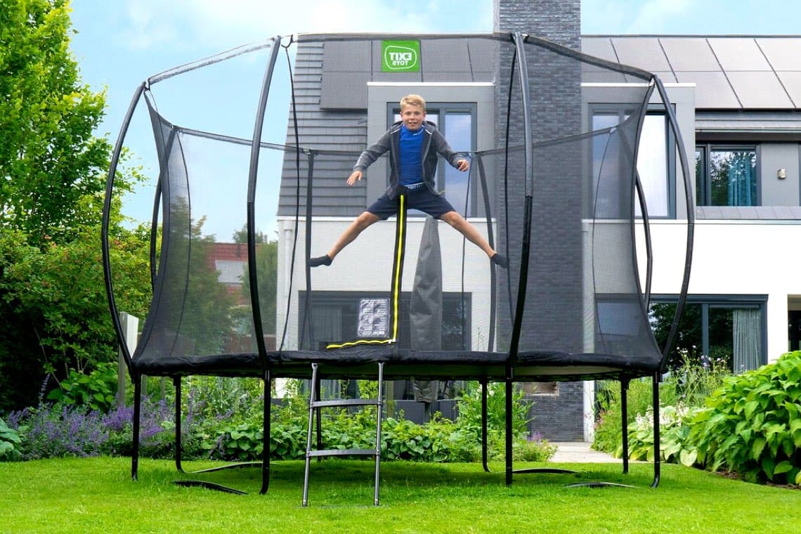 Junge springt mit gespreizten Beinen auf rundem Trampolin mit Netz im Garten vor einem modernen Haus