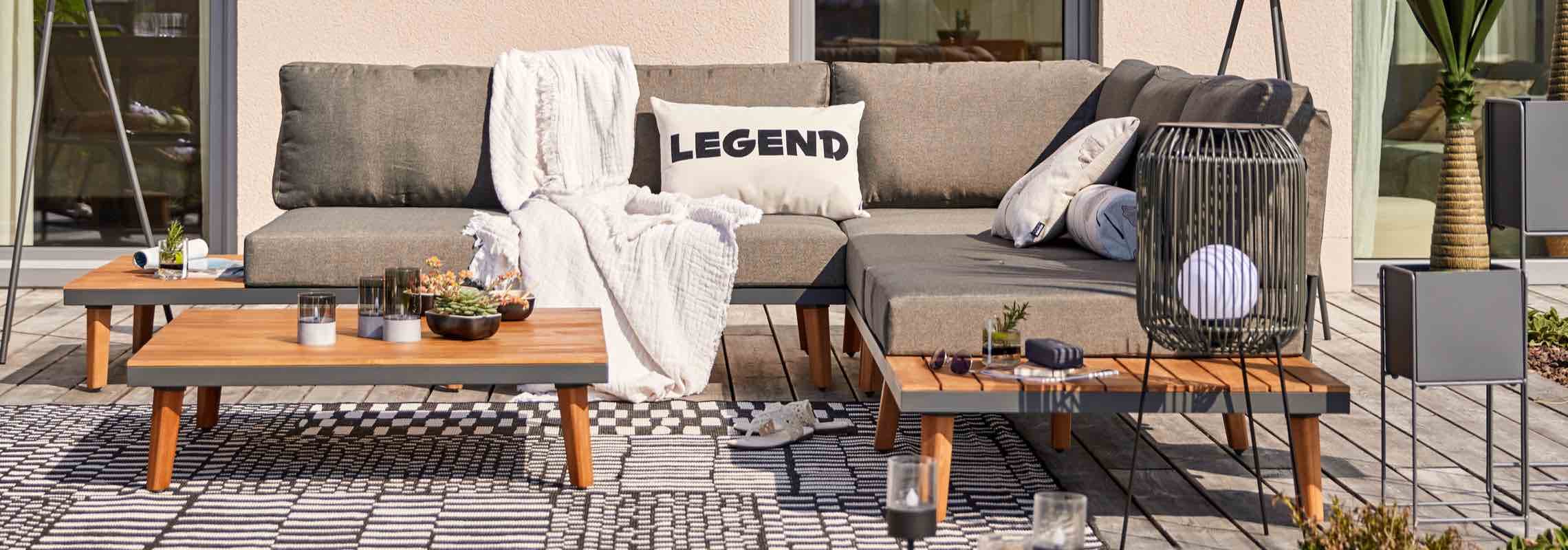 Kissen mit Aufdruck LEGEND auf Ecksofa, davor zwei niedrige Tische mit Deko auf Terrasse mit Teppich