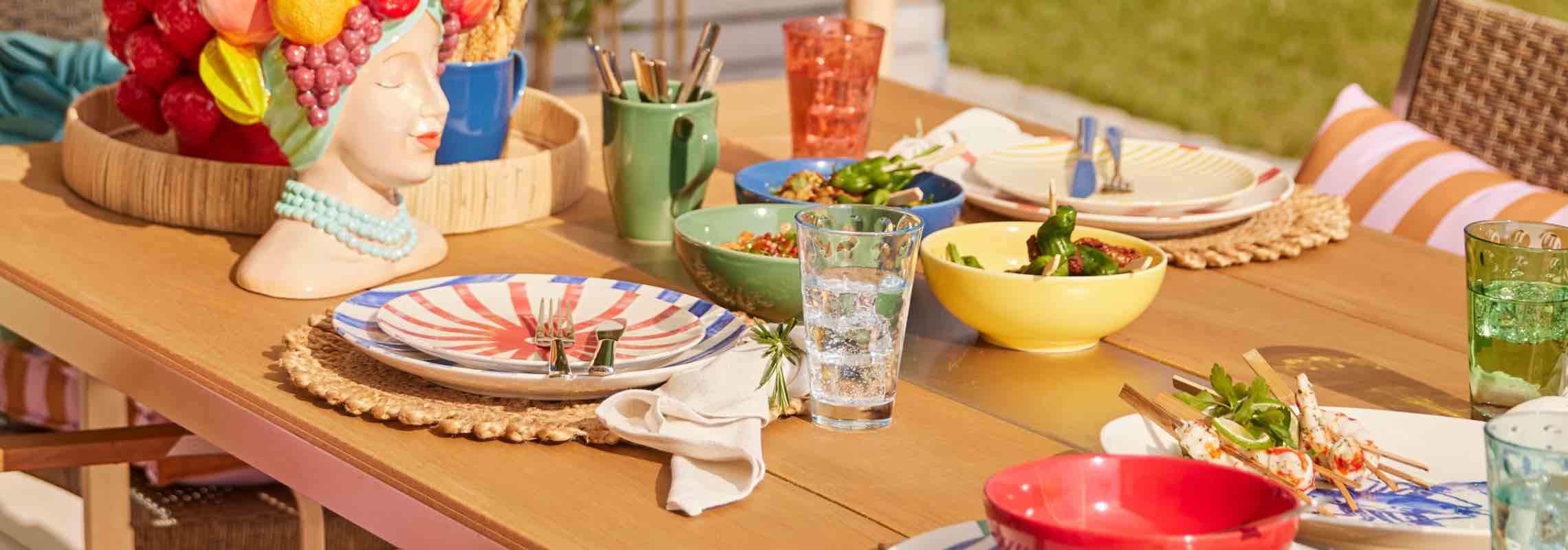 Gedeckter Gartentisch mit bunten Tellern, Schalen, Gläsern und kleinen Speisen auf einem langen Holztisch