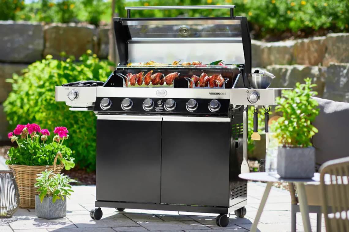 Großer Gasgrill im Garten, Deckel geöffnet, mit mehreren Spießen Fleisch und Gemüse auf der Grillfläche