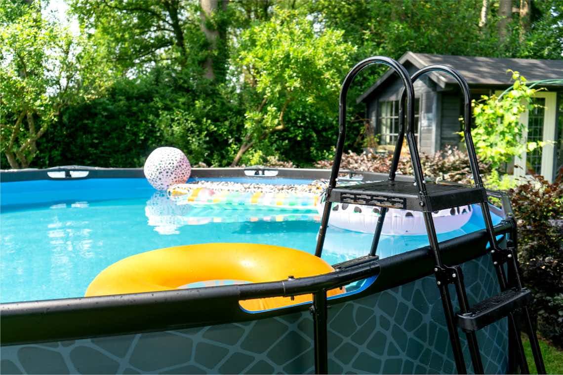 Runder Aufstellpool im Garten mit Leiter, gelbem Schwimmring und bunten Luftmatratzen im Wasser