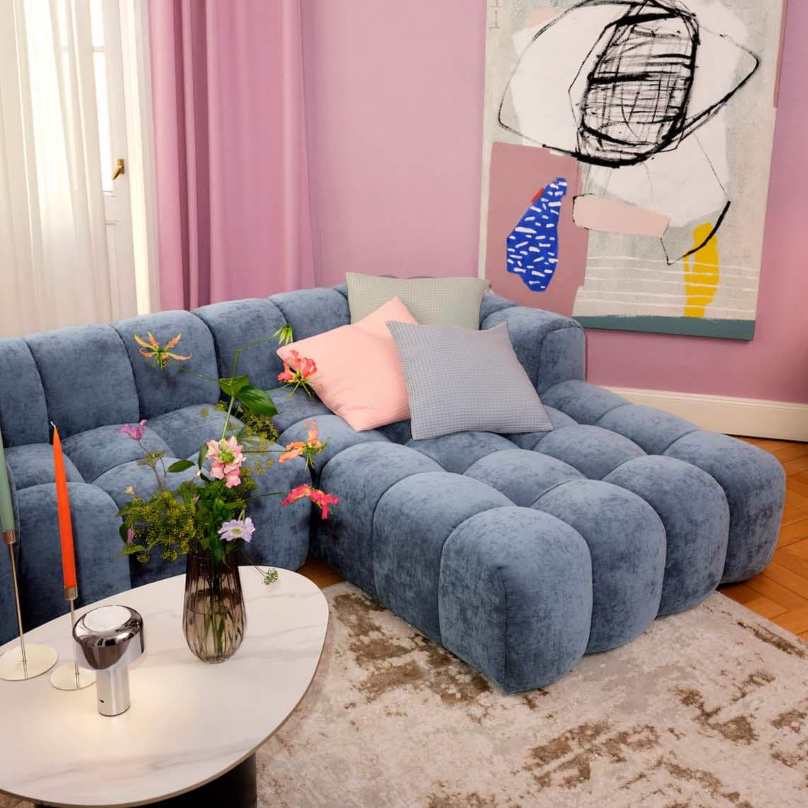 Blaue modulare Wohnlandschaft mit pastellfarbenen Kissen neben Couchtisch mit Blumenstrauß und Kerzen in rosa Wohnzimmer