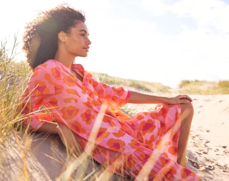 Frau in pink-orange gemustertem Kleid sitzt entspannt im Sand einer sonnigen Dünenlandschaft