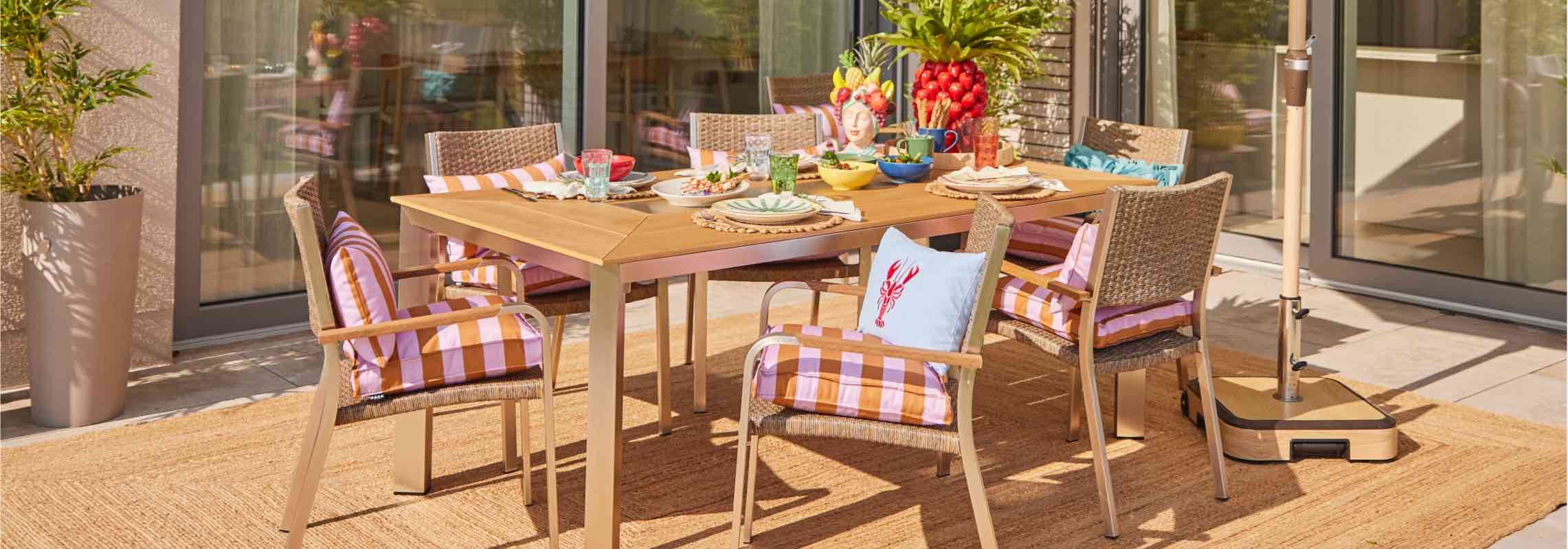Gedeckter Gartentisch mit buntem Geschirr, Obstschale und sechs Stühlen mit gestreiften Kissen auf Terrasse
