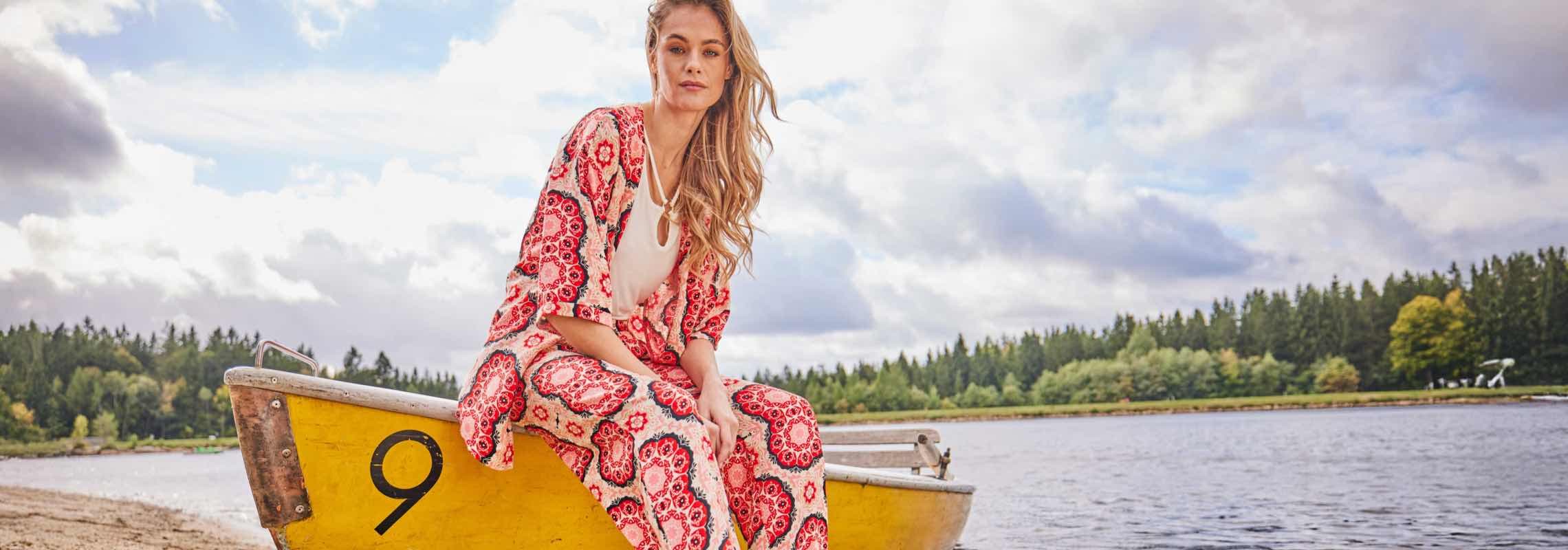 Frau im gemusterten Zweiteiler sitzt auf einem gelben Ruderboot am See vor bewaldetem Ufer und bewölktem Himmel