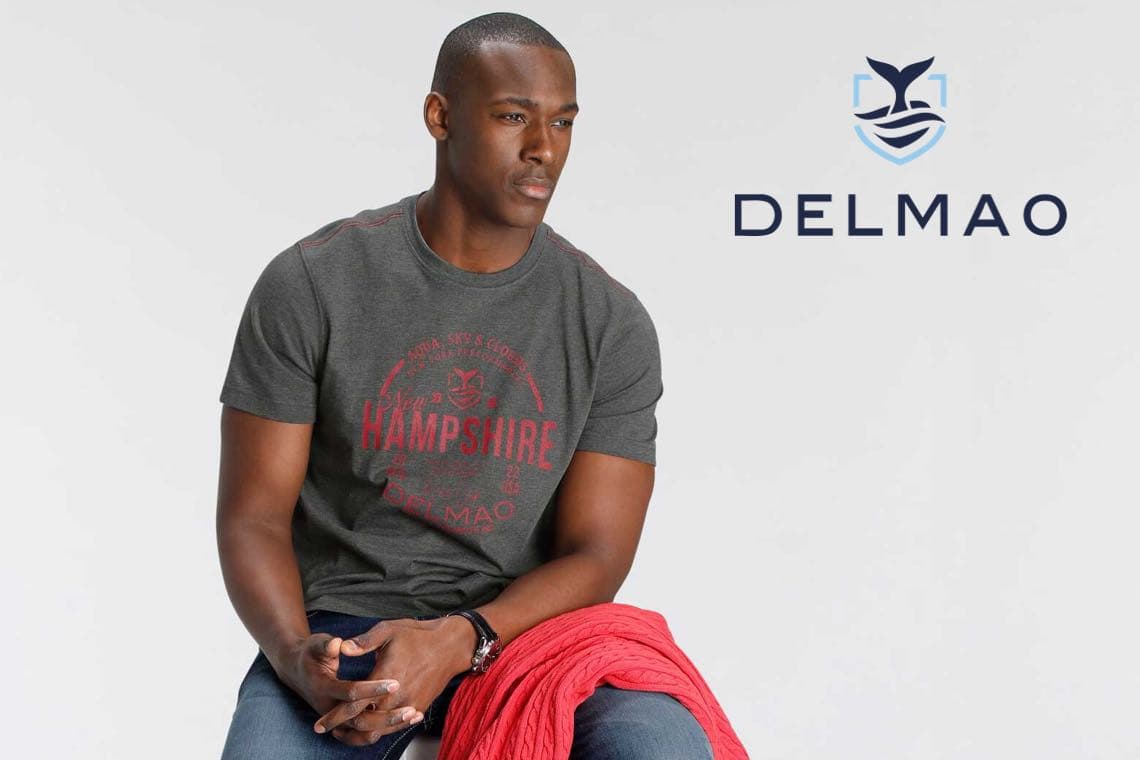 DELMAO Logo rechts – Mann sitzt und trägt graues T‑Shirt mit rotem Hampshire‑Print und rotem Pullover auf dem Schoß