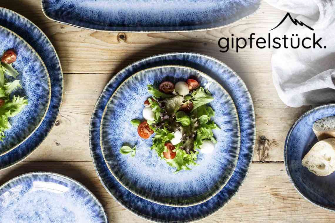 gipfelstück – blauer Teller mit grünem Salat, Tomaten und Mozzarella auf Holztisch mit weiteren Tellern und Brot