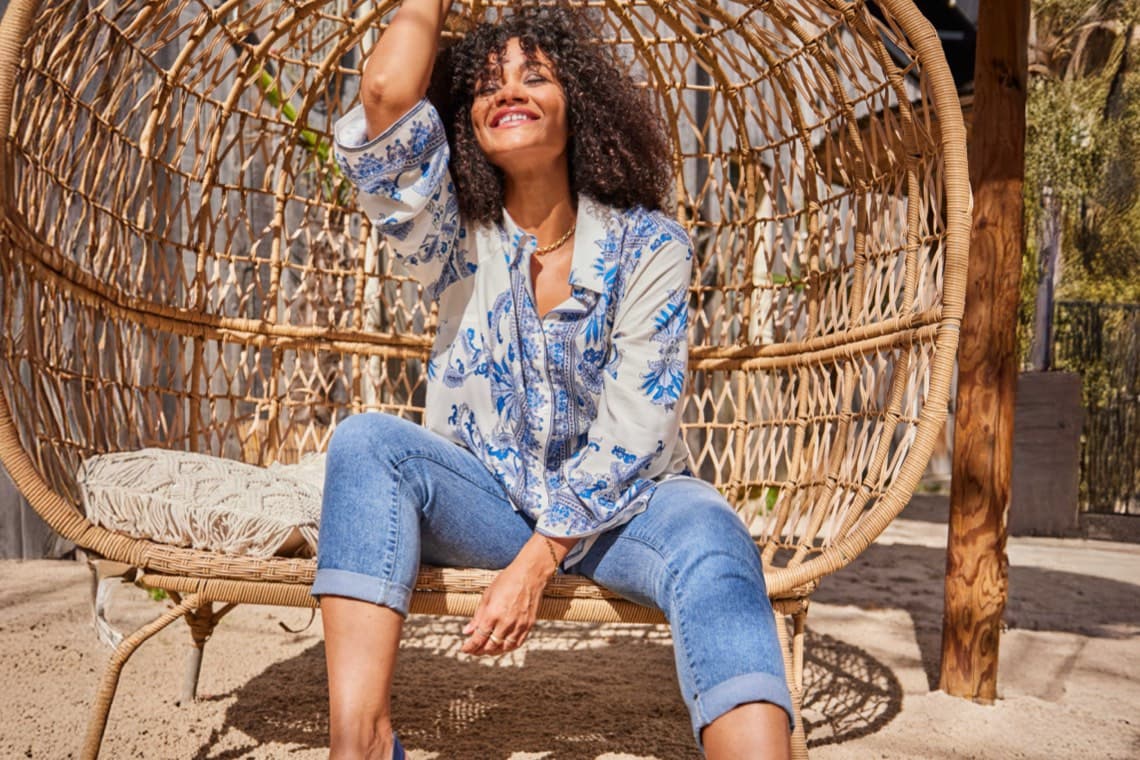 Frau in gemusterter Bluse und gekrempelter Jeans sitzt entspannt in einem großen Hängesessel aus Flechtwerk draußen