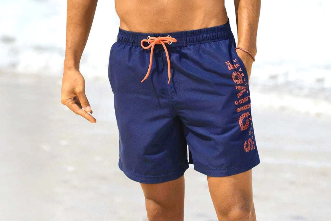 Mann am Strand trägt dunkelblaue Badeshorts mit orangener Kordel und Schriftzug am Bein