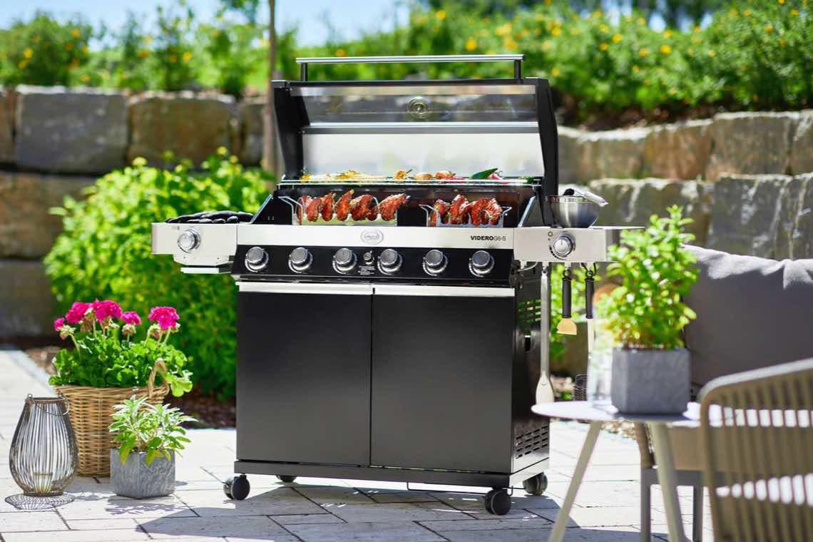 Großer Grillwagen im Garten, Deckel offen, Fleischspieße und Gemüse auf dem Rost, umgeben von Blumentöpfen