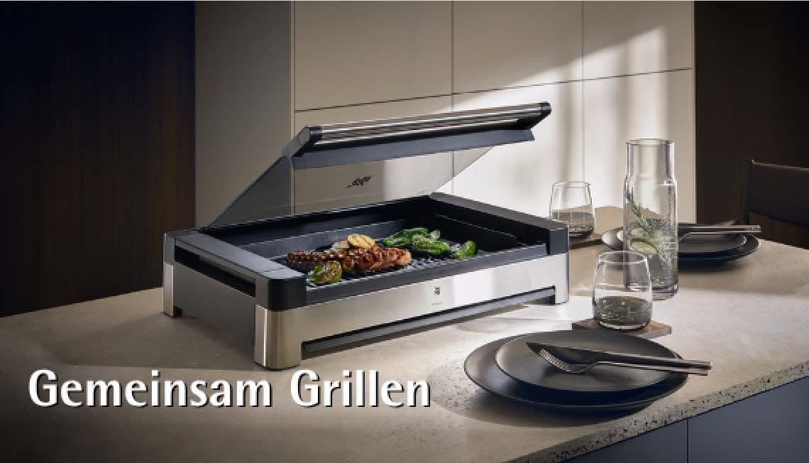 Gemeinsam Grillen – Tischgrill mit offenem Deckel, Gemüse und Fleisch, gedeckter Esstisch mit Tellern und Gläsern