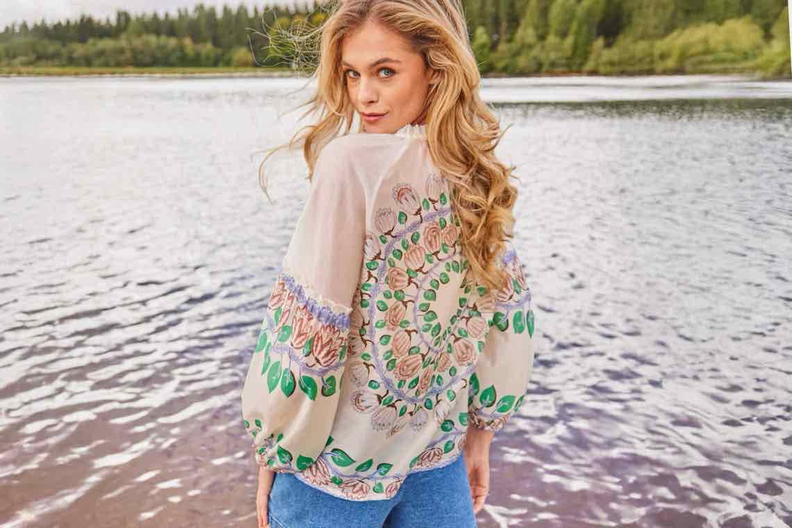 Frau mit langen Locken steht am See, trägt helle Bluse mit buntem Blumenmuster und blaue Hose, Rücken zur Kamera