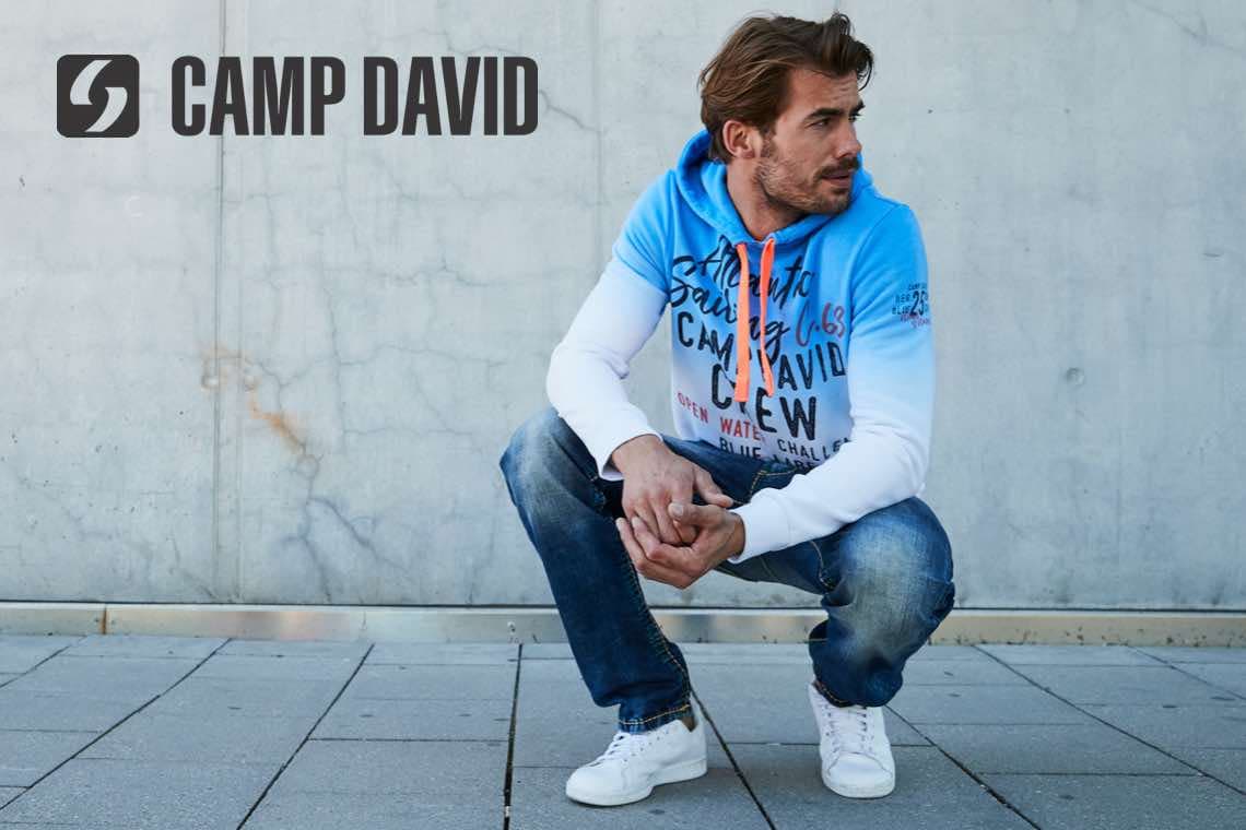 CAMP DAVID – Mann in blauem Hoodie und Jeans hockt vor Betonwand auf gepflastertem Boden