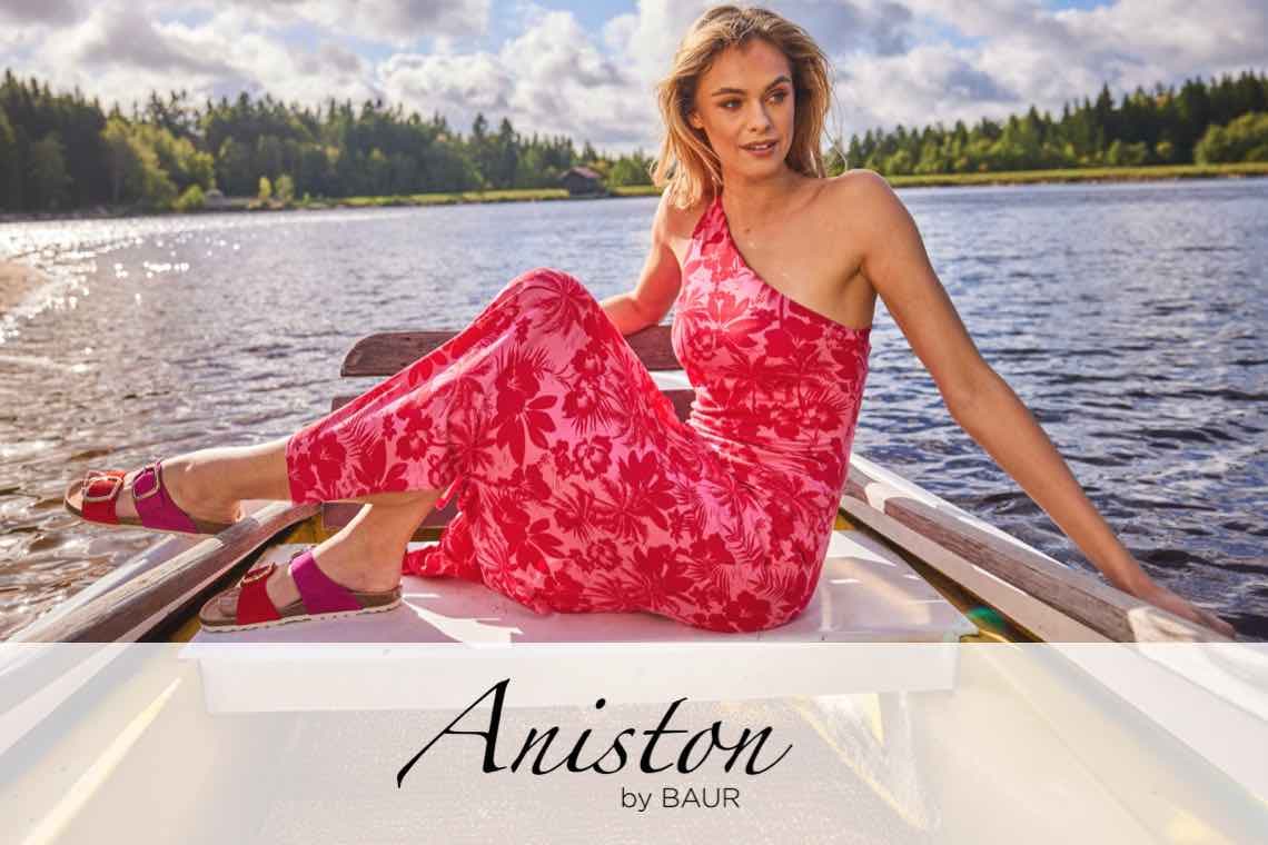 Aniston by BAUR – Frau im roten Ein-Schulter-Kleid sitzt auf einem Boot auf einem See vor bewaldetem Ufer