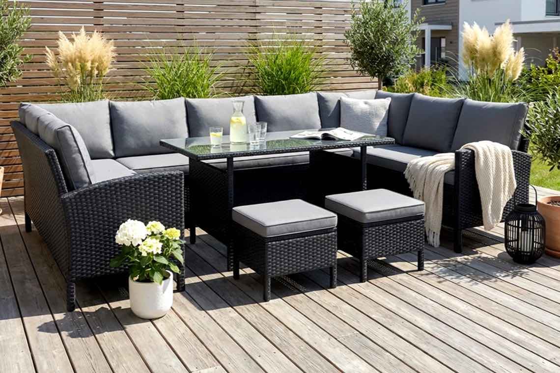 Eckige Gartenlounge mit grauen Polstern, Tisch mit Karaffe und Gläsern, Hocker und Decke auf Holzterrasse vor Sichtschutz
