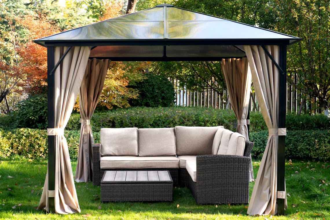 Gartenpavillon mit offenen Vorhängen, darunter Ecksofa und niedriger Tisch auf Rasen vor Hecke