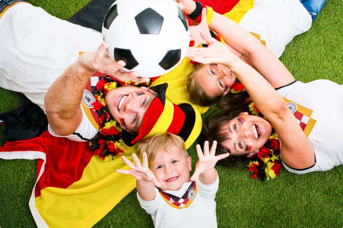 Fans in Deutschlandfarben liegen im Kreis auf dem Rasen und strecken gemeinsam die Hände nach einem Fußball aus
