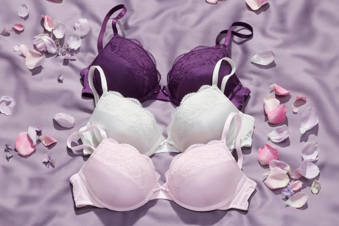 Drei Spitzen-BHs in Lila, Weiß und Rosa liegen auf violettem Stoff, umgeben von verstreuten Blütenblättern