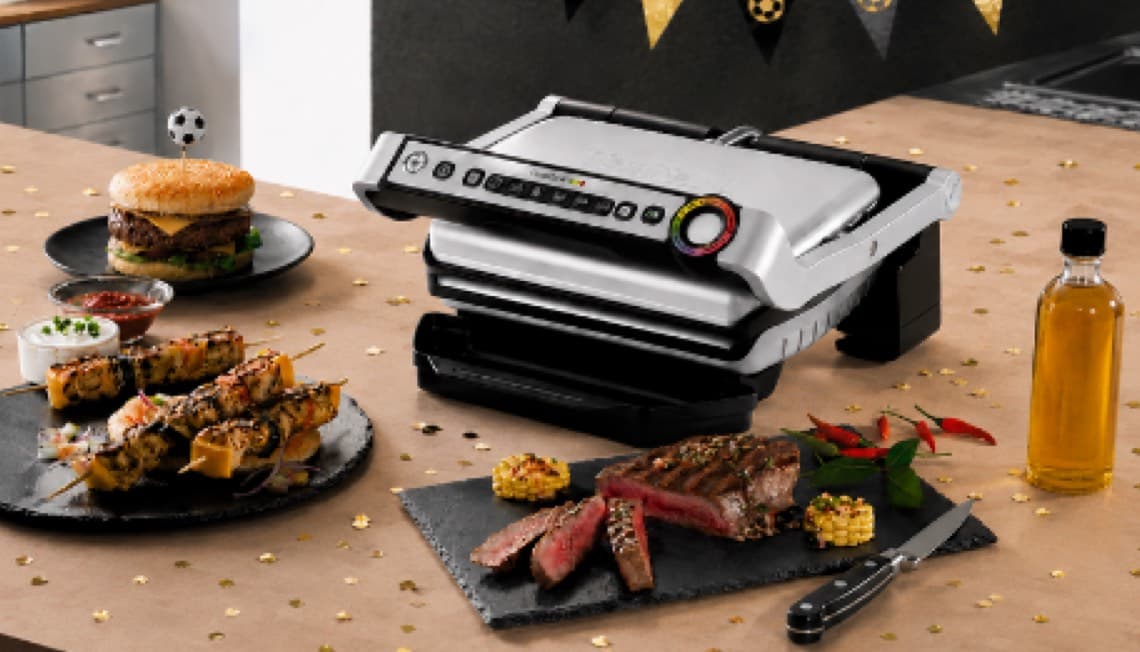 Kontaktgrill auf Arbeitsplatte, umgeben von Burger, Fleischspießen, Steak, Mais, Soßen und Flasche mit Öl
