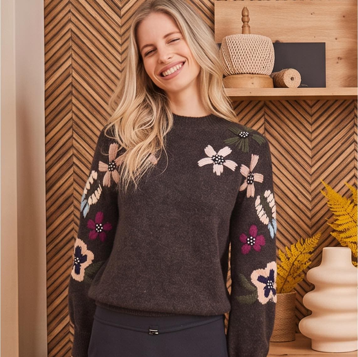 Frau mit langem Haar trägt dunklen Pullover mit bunten Blumenärmeln, steht vor Holzregal mit Dekoobjekten