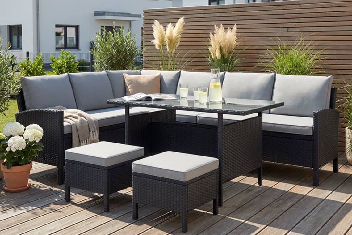 Eckige Gartenlounge mit grauen Polstern, Glastisch mit Karaffe Limonade und Gläsern auf Holzterrasse vor Sichtschutzwand