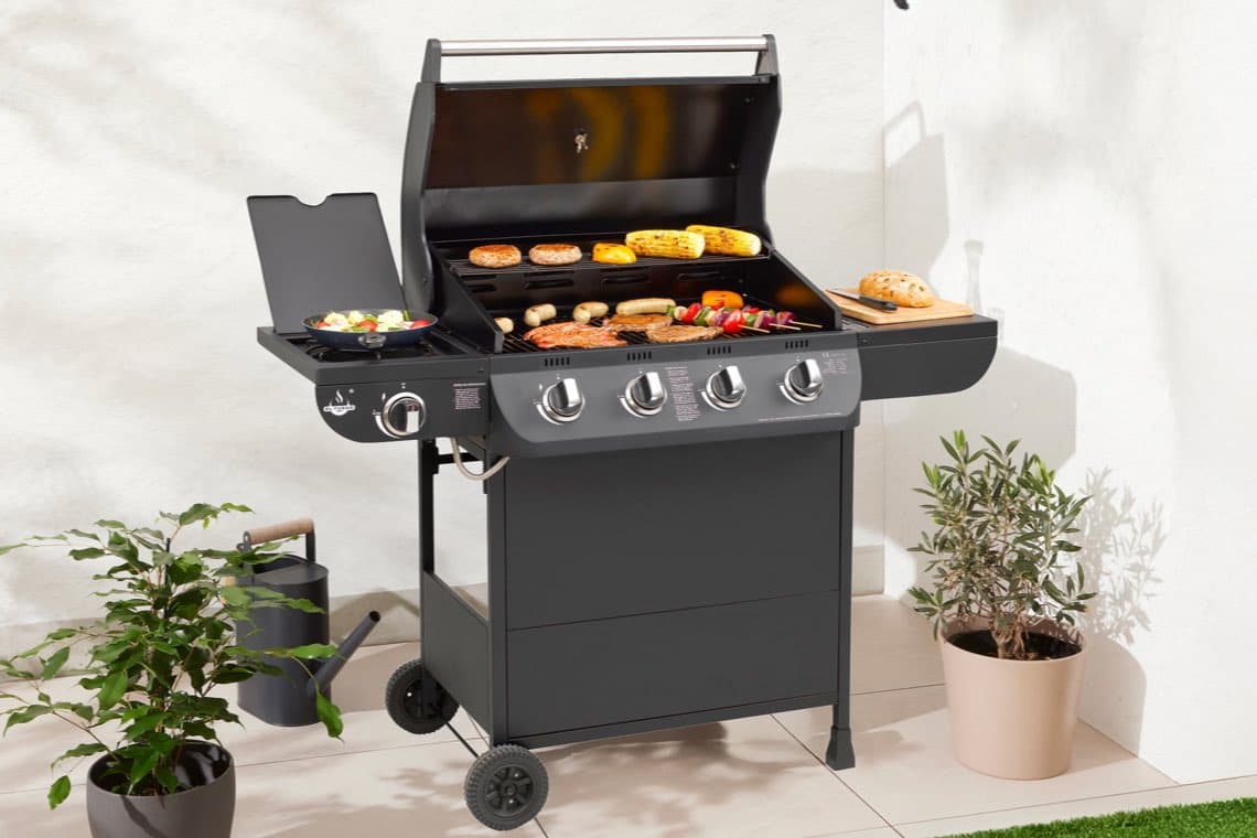 Gasgrill mit vier Drehknöpfen, aufgeklappt, mit Fleisch, Gemüse, Maiskolben und Pfanne mit Gemüse auf Terrasse