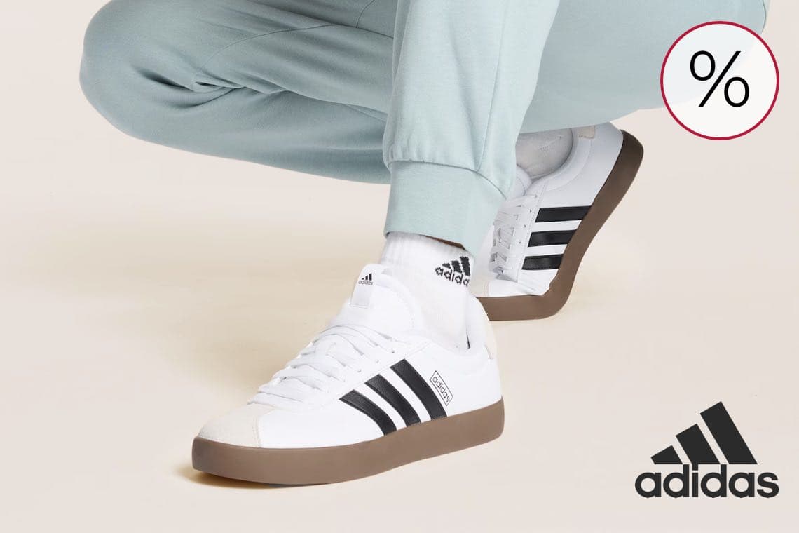 %-Zeichen Angebot – Person in hellgrüner Hose kniet, trägt weiße adidas Sneaker mit schwarzen Streifen und Logo