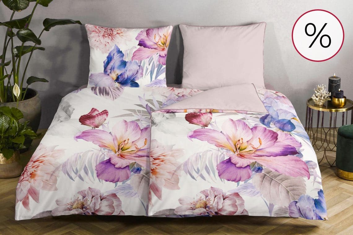 Prozentzeichen-Angebot – Bett mit geblümter Bettwäsche in Rosa und Lila, daneben Pflanze und kleiner Beistelltisch