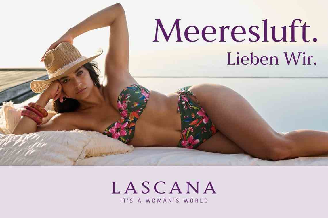 Meeresluft. Lieben wir. Frau im Blumenbikini liegt mit Strohhut entspannt auf Kissen am Wasser, Lascana Logo unten