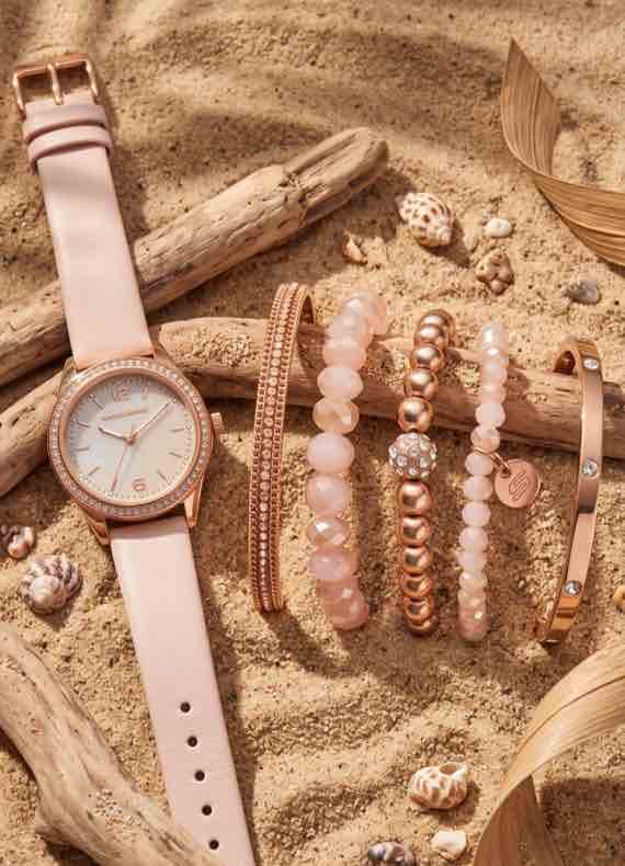 Armbanduhr und mehrere Armbänder in Rosa und Gold liegen dekorativ im Sand zwischen Treibholz und kleinen Muscheln