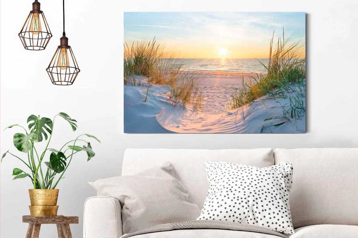 Wohnzimmer mit Sofa, Punktkissen, Pflanze und Wandbild eines Sonnenuntergangs über sandigem Strand mit Dünengras