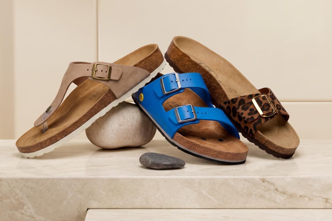 Drei Sandalen auf Steinpodest: beige Zehenstegsandale, blaue Sandale mit zwei Riemen, braune Sandale mit Leopardenmuster