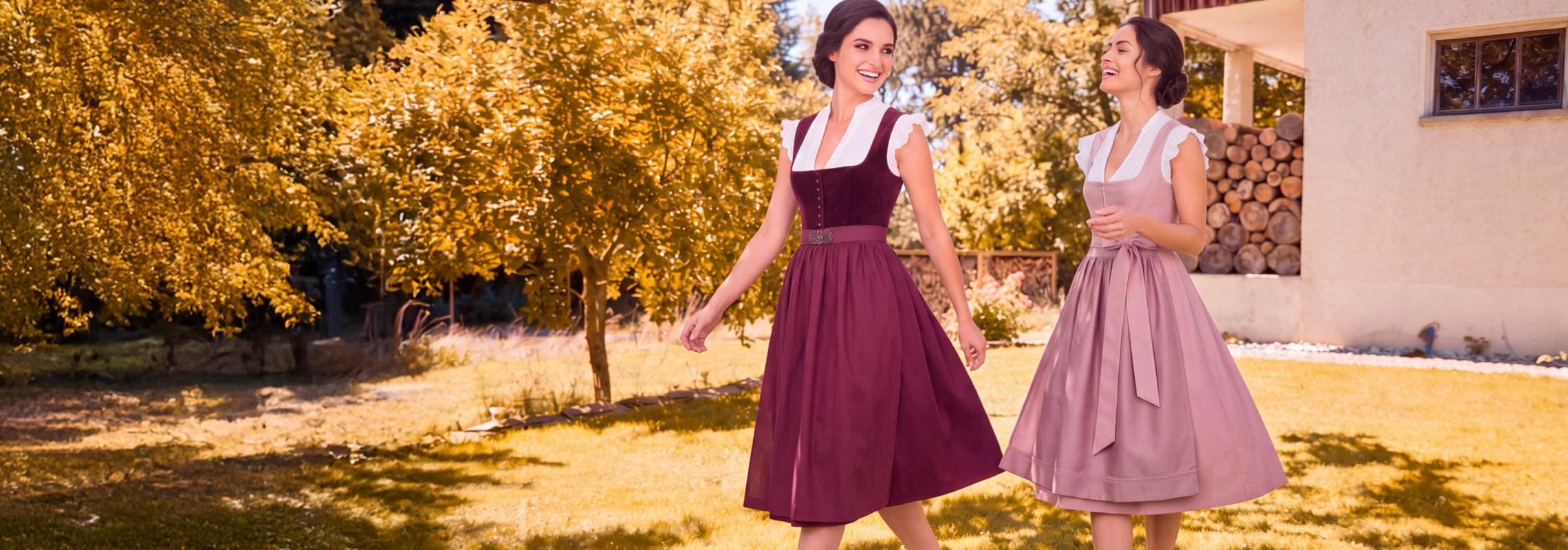 Zwei Frauen in Dirndln gehen lachend über eine Wiese vor Haus und Bäumen