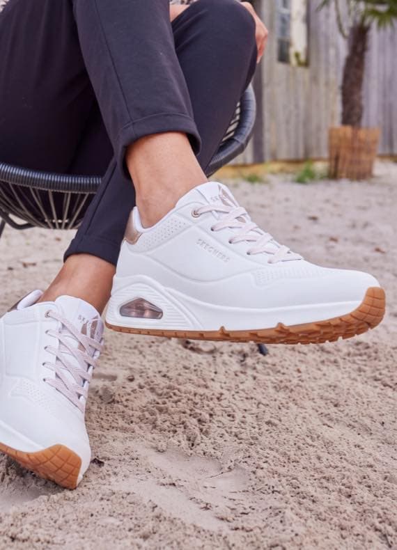 Person sitzt am Strand, zeigt weiße Sneaker mit heller Sohle und Schnürung, Beine in dunkler Hose entspannt ausgestreckt