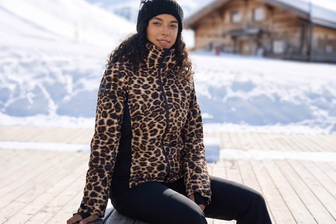 Frau in Leopardenjacke und Mütze sitzt auf Holzbank vor verschneiter Berghütte