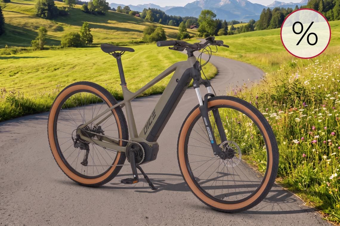 Prozentzeichen für Angebot – E-Bike steht auf Landstraße vor grünen Wiesen und Bergen