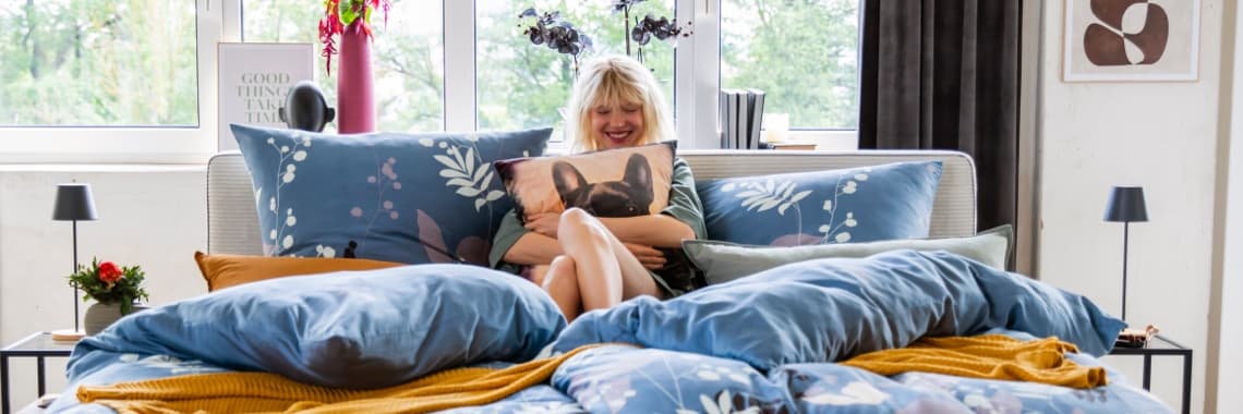 Ein gemütliches Schlafzimmer mit einem großen Bett, das mit bunten, floral gemusterten Bettwäsche und Kissen dekoriert ist. Auf dem Bett liegen auch eine gelbe Decke und ein Kissen mit einem Hundemotiv. Im Hintergrund sind Fenster mit Blick ins Freie.