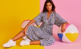Eine Person sitzt in einem gestreiften, lässigen Kleid und trägt weiße Sneakers. Der Hintergrund ist in zwei lebhaften Farben unterteilt, Gelb und Pink, und es sind bunte Strandbälle zu sehen, die eine sommerliche Atmosphäre schaffen.