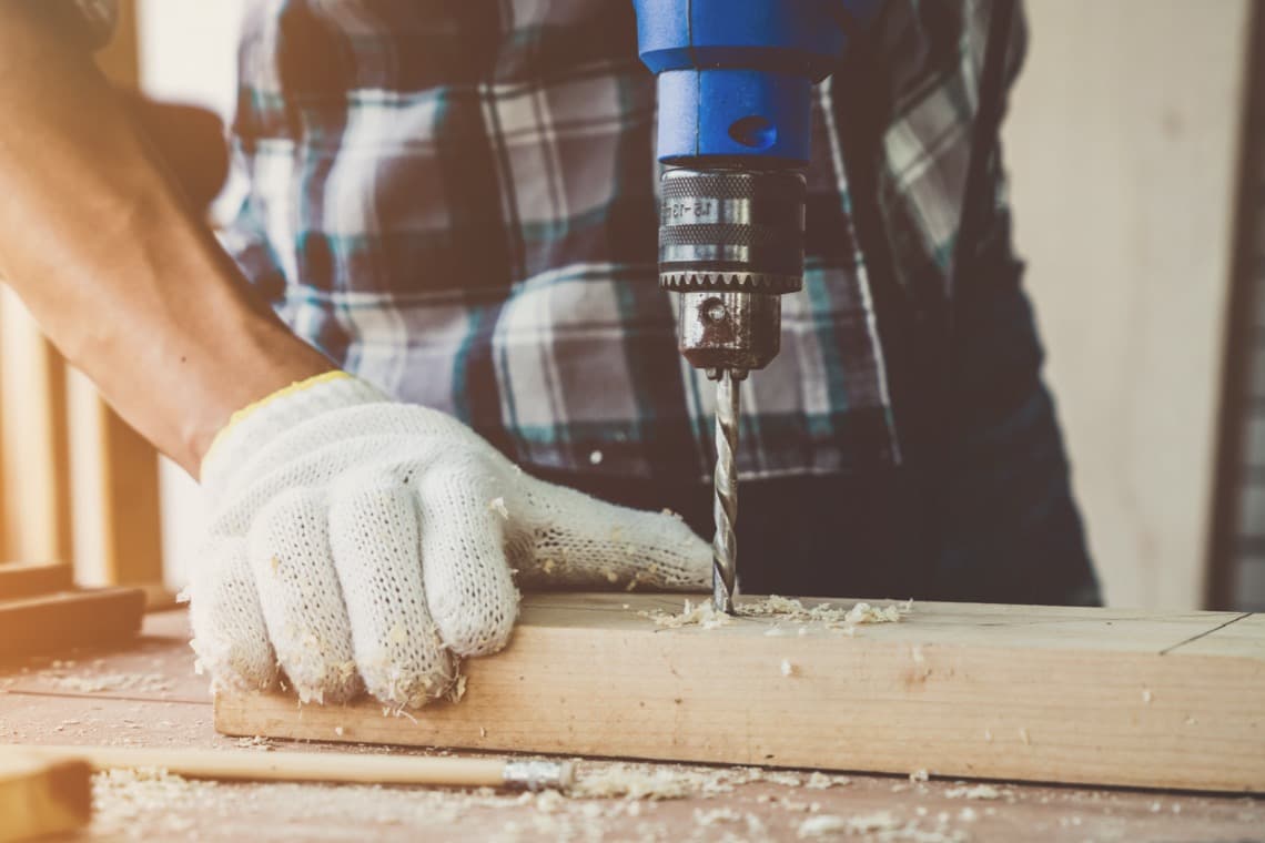 Ein Mann in einem karierten Hemd bohrt mit einem blauen Bohrer in ein Holzstück, das auf einem Tisch liegt. Er trägt einen grauen Arbeitshandschuh.