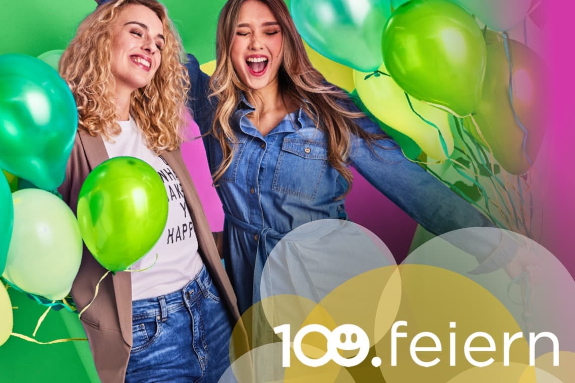 Zwei Frauen stehen vor einem bunten Hintergrund mit grünen und goldenen Luftballons. Sie lächeln und posieren fröhlich für die Kamera.