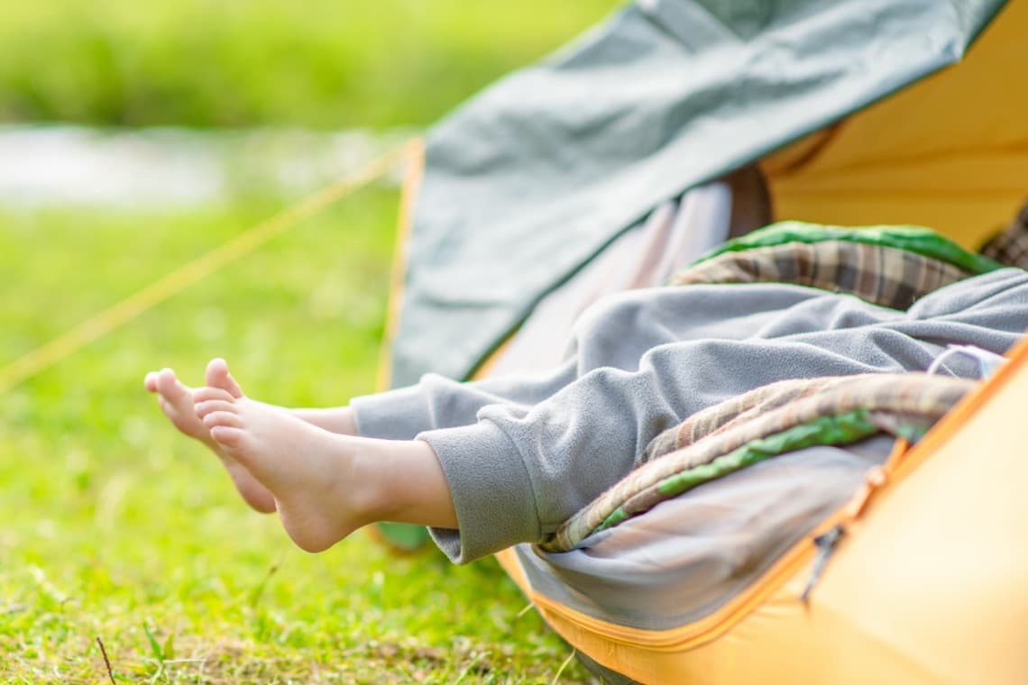 Kinderfüße ragen aus einem orangefarbenen Zelt, umgeben von grüner Wiese. Der Schlafsack ist aufgeschlagen und zeigt eine gemütliche Innenausstattung.