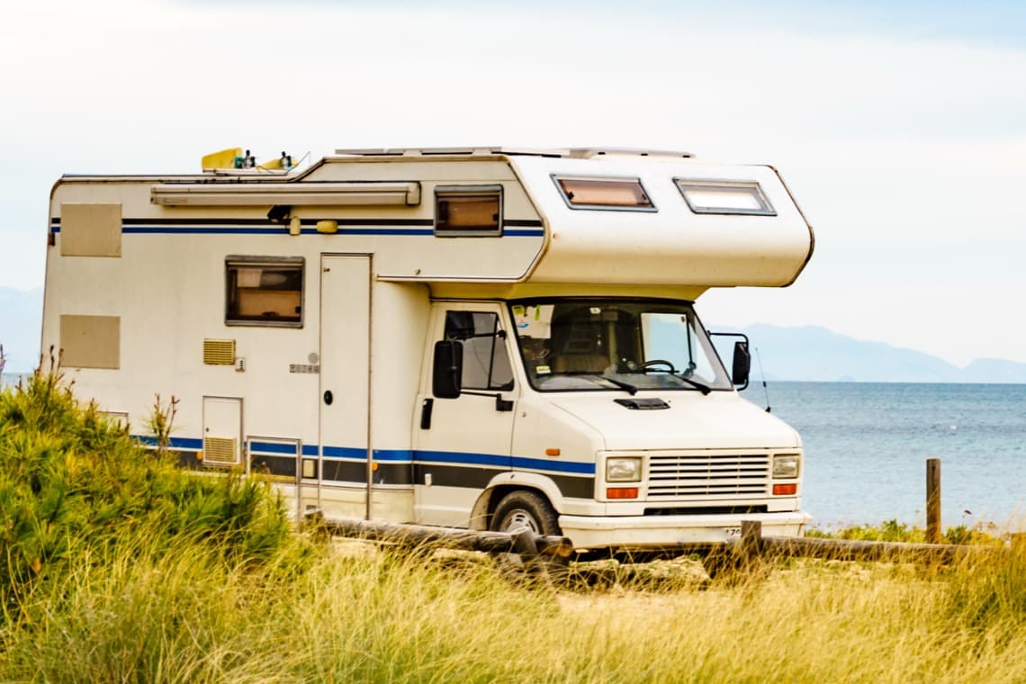 Ein weißes Wohnmobil mit blauen Streifen steht am Strand. Im Hintergrund ist das Meer und der Himmel zu sehen, umgeben von Gras.
