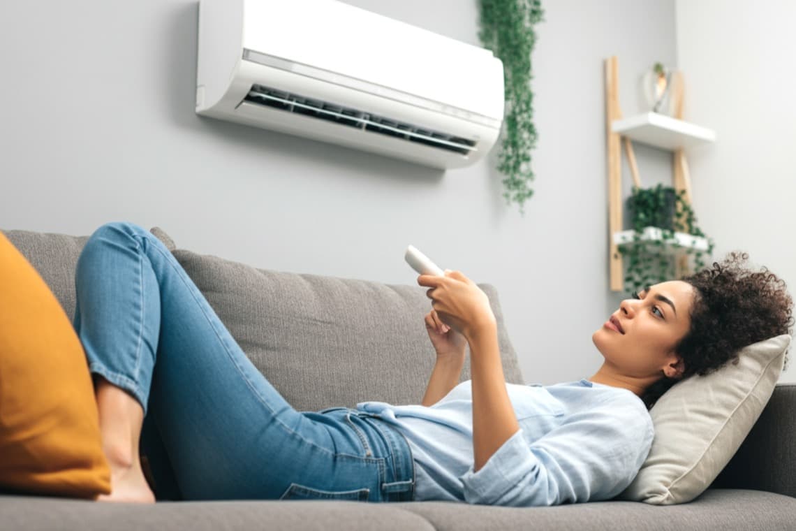 Eine Frau liegt auf einem grauen Sofa und hält eine Fernbedienung in der Hand. Über dem Sofa hängt eine Klimaanlage. Der Hintergrund ist hell.