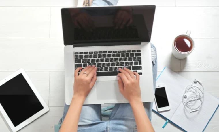 Person tippt auf Laptop, daneben Tablet, Notizblock, Smartphone, Kopfhörer und Tasse Kaffee auf hellem Boden.
