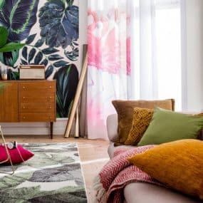 Wohnzimmer mit bunter Wandtapete, Sofa mit bunten Kissen und Holzmöbeln, helle Vorhänge mit rosa Flamingos.